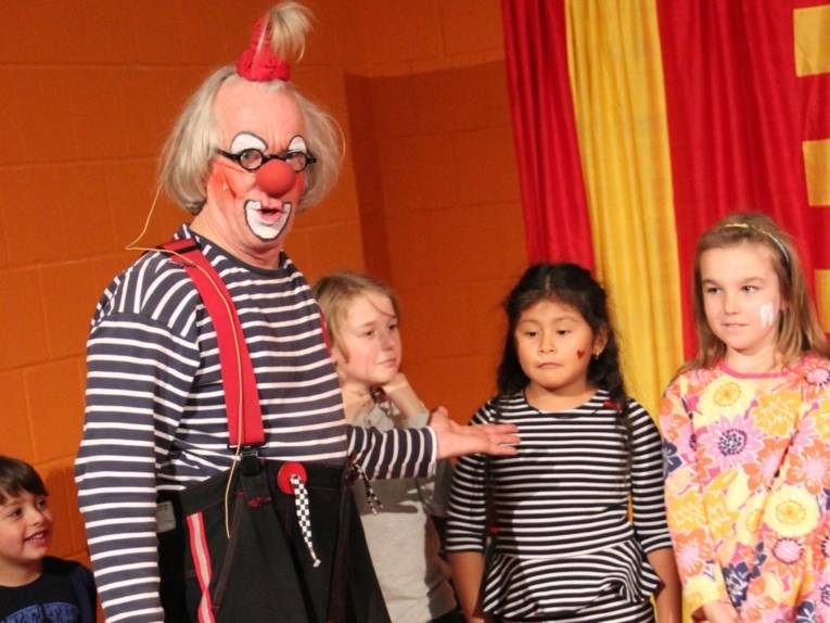 Ein Clown deutet mit der linken Hand auf drei Mädchen, die neben ihm stehen.