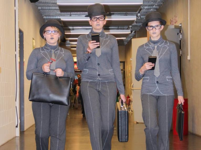 Drei junge Frauen sind als graue Herren verkleidet und laufen im Gleichschritt einen Gang entlang. Sie tragen Melone, eine Aktentasche und Taschenrechner. Ihre Anzüge sind mit weißer Textilfarbe auf grauen Trikotagen stilisiert dargestellt. 