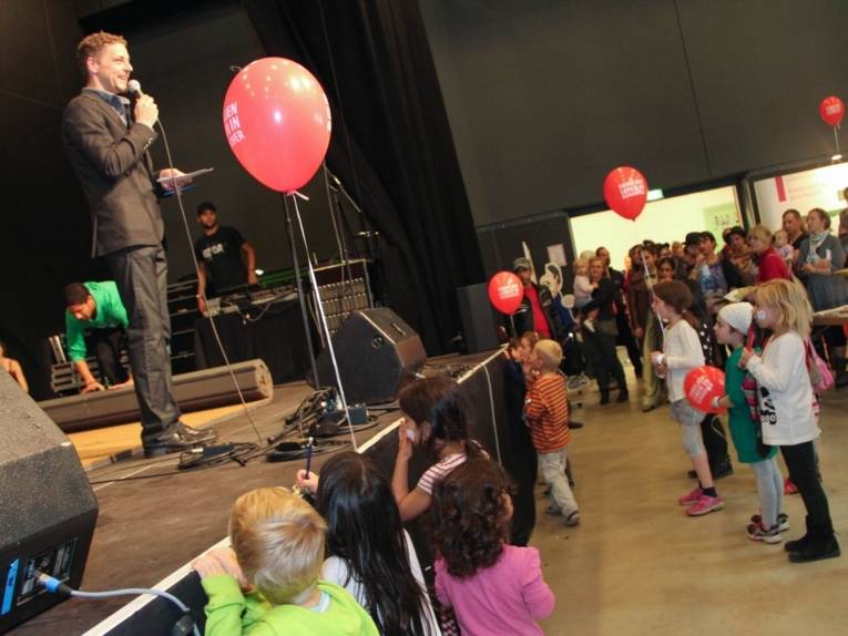 Auf einer Bühne steht ein Mann mit Mikrofon, davor im Zuschauerraum stehen Kinder mit Luftballons und Erwachsene.