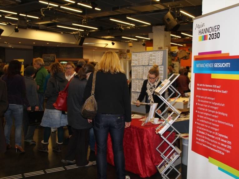 Anmeldetresen mit Namensschildern für die Tagung "Zweites Forum Familie" mit zahrliechen Gästen im Hintergrund. Vorn rechts im Bild ist ein Aufsteller mit dem Schriftzug "Mein Hannover 2030" und der Überschrift "Mitmachende gesucht" zu sehen.