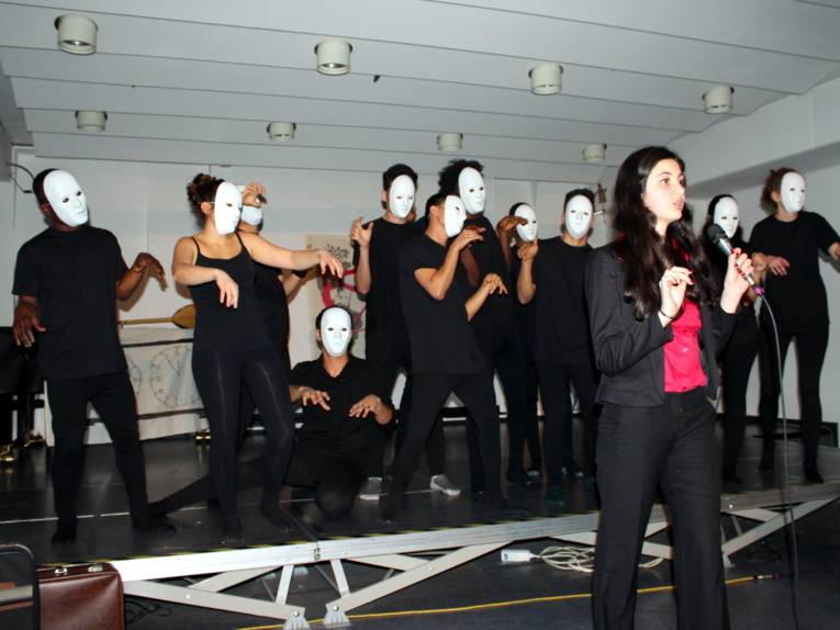 Eine Jugendliche steht vor der Bühne des VHS-Saals und spricht in ein Mikrofon. Hinter ihr stehen etwa 12 weitere Personen, die allesamt neutrale Masken vor dem Gesicht tragen und ihre Arme nach vorne strecken.