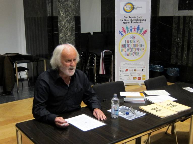 Ein Mann sitzt hinter einem Tisch und spricht. Auf seinem Tisch liegen mitgebrachte Bücher und Aufsätze und eine kleine durchsichtige Plastikflasche mit Wasser. Rechts hinter ihm ist ein Roll-Up des Runden Tisch für Gleichberechtigung und gegen Rassismus mit seinem typischen Logo, in dem sich Strichmännchen verschiedener Farben und Größen an den Händen anfassen.
