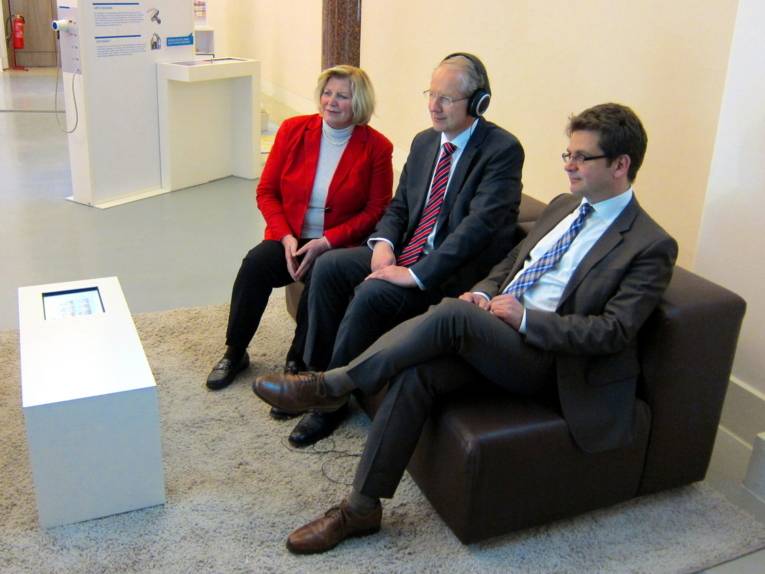 Drei Personen sitzen nebeneinander auf einem Sofa. Die mittlere person hat ein Kopfhörerpaar mit Bügel auf dem Kopf. Alle drei blicken nach vorne.