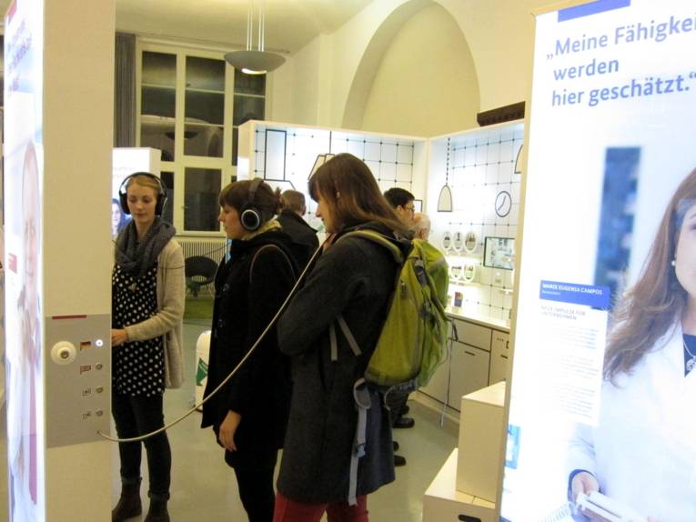 Drei Frauen stehen an einer multimedialen Ausstellungswand. Zwei davon haben Kopfhörer aufgesetzt, die mit der Stellwand verbunden sind. Eine weitere Frau hält ein Telefon ans Ohr, das ebenfalls mit der Wand verbunden ist. Im Intergrund sind weitere Stellwände erkennbar, an denen weitere personen von hinten erkennbar sind. Sie beschäftigen sich mit der Ausstellungswand vor sich.