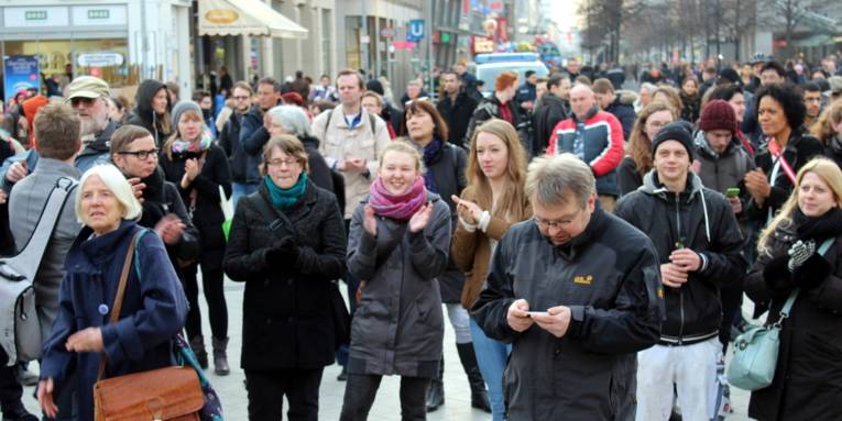 Blick von der Bühne neben der Uhr am Kroepcke in Richtung Steintor: Etwa 100 Teilnehmer/innen blicken zur Bühne in Richtung Opernplatz.