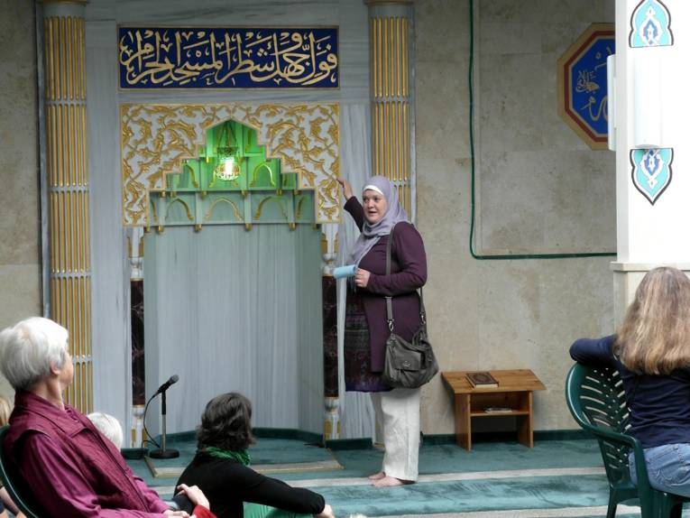 Die Referentin steht vor einer Einbuchtung, die in die Gebetsrichtung (Qibla) zeigt. Über der Nische ist das muslimische Glaubensbekenntnis (Schahada) in arabischer Schrift. Links und rechts davon sind Marmorsäulen.
