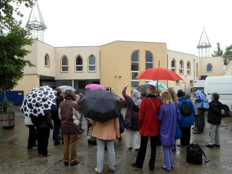 Etwa 15 Personen stehen mti Regenschirmen vor der Moschee. Links und rechts von Gebäude ragen gläserne Minarette in die Luft.