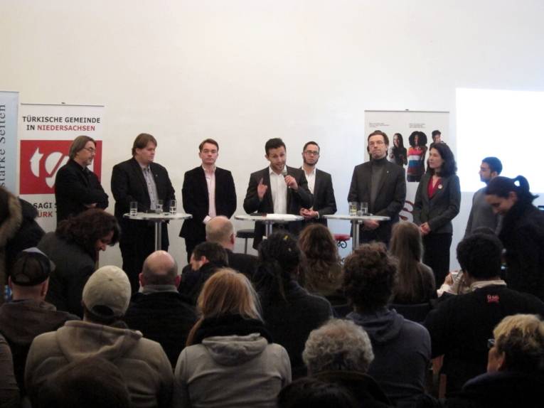 Acht Personen stehen vor weißen Stehtischen. Der Mittlere hält ein Mikrofon in der Hand und spricht. Unten im Bild sind Zuschauer/innen zu erkennen.