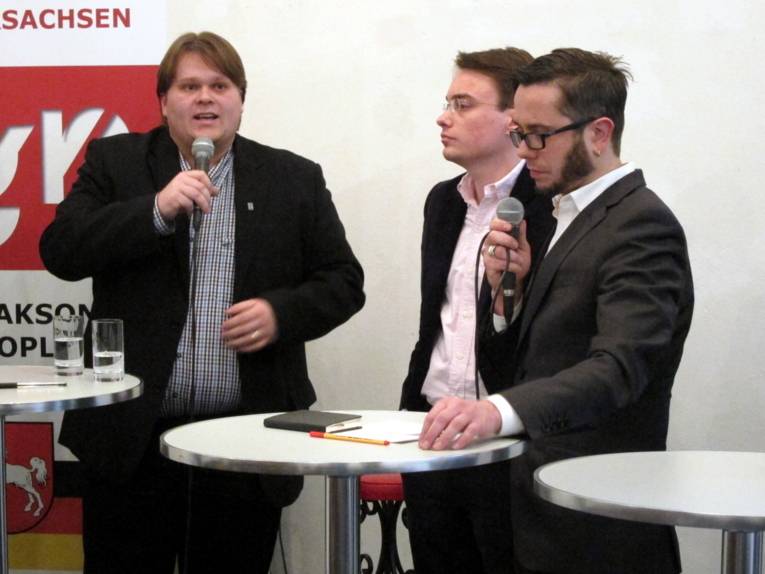 Drei Personen stehen hinter zwei Stehtischen. Zwei davon halten ein Mikrofon in der Hand und die linke Person spricht.