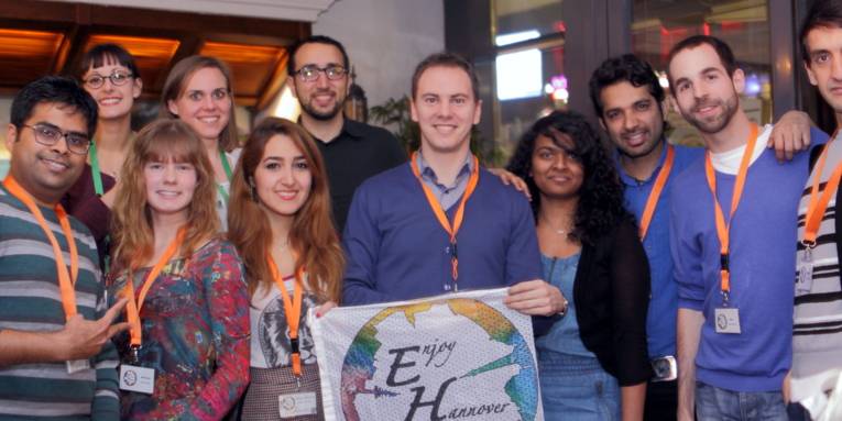 Elf Personen stehen nebeneinander in zwei Reihen und halten vor sich ein Stofftransparent mit dem Aufdruck "Enjoy Hannover" auf.