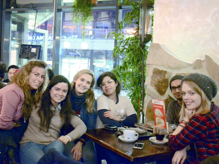 Sechs gut gelaunt aussehende Personen - fünf Frauen und ein Mann - sitzen um einen Tisch in einem Café und blicken in die Kamera.