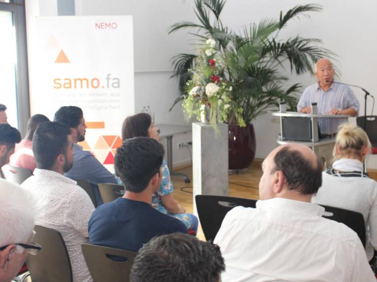 Ein mann steht hinter einem Redner*innenpult uns spricht. Vor ihm sitzen knapp 15 Menschen in Stuhlreihen und blicken nach vorn zum Sprecher.