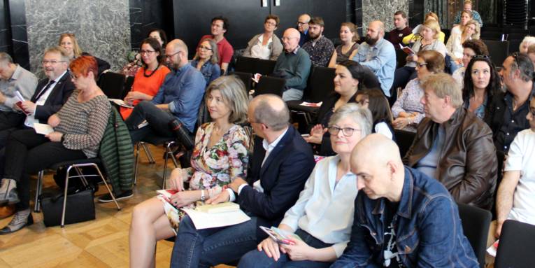 Etwa 20 Personen sitzen in Stuhlreihen im Mosaiksaal des Neuen Rathauses und blicken nach vorn.