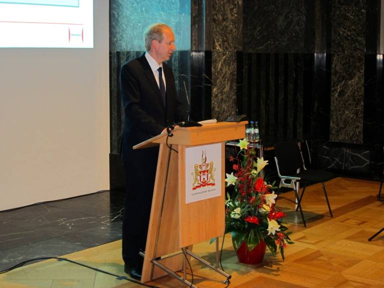 Ein Mann steht hinter einem Rednerpult und trägt vor. Das Pult trägt das Hannover-Wappen, rechts daneben steht ein großes Blumengesteck. An der Wand hinter dem Redner läuft eine Beamerpräsentation.