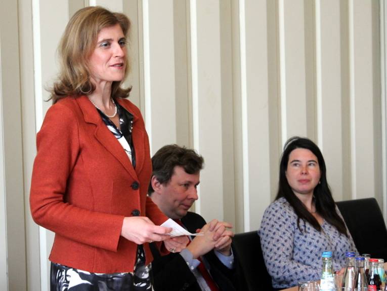 Eine Frau (links) steht hinter einem Tisch und spricht im Gobelinsaal. Rechts neben ihr sitzen ein Mann und eine Frau