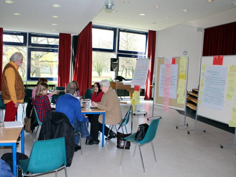 Ein sechsköpfige Gruppe sitzt um einen Tisch. Der Moderator steht links daneben.