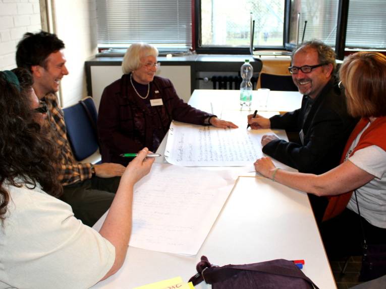 Fünf Personen, drei Frauen und zwei Männer, sitzen um einem Tisch, auf dem Stifte und Papiere liegen. Einer der Männer beschreibt ein großes Blatt Papier mit einem großen Stift.