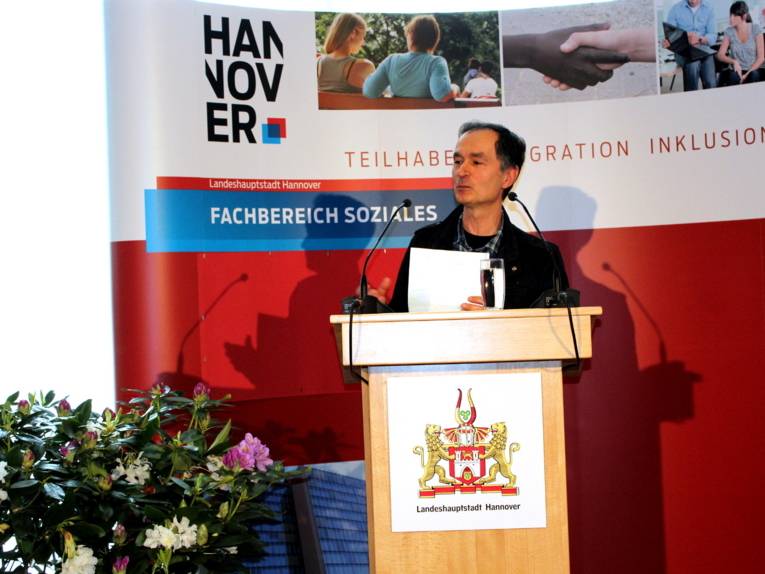 Ein Mann steht hinter einem Pult und spricht in ein Mikrofon. Dabei hält er eien dünnen Stapel Papieren in der Hand.