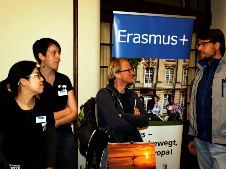Vier Personen stehen vor einem Stand, auf dem "Erasmus" steht und sprechen miteinander.