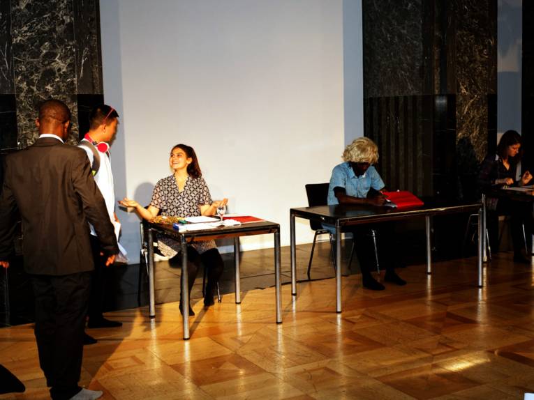 Zwei Personen stehen vor einem "Sachbearbeiter-Tisch", an dem eine weitere Darstellerin sitzt. Rechts daneben sitzen zwei weitere Darsteller/innen an Tischen und blicken auf 