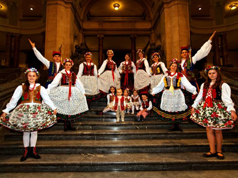Ein Dutzend Mitglieder der lokalen Folkloregruppen „Polonia“ und „Lajkonik“ präsentieren ihre farbenprächtigen Trachten auf der steinernen Treppe in der Rathaushalle