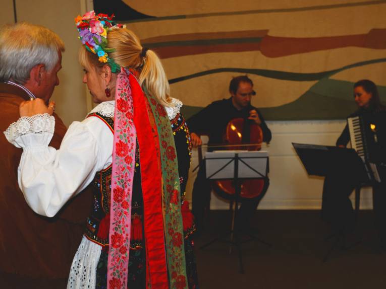 Ein Mann und eine Frau in Trachtenkleidung unterhalten sich, während ein Cello-Spieler und eine Akkordeon-Musikerin im Hintergrund aufspielen.
