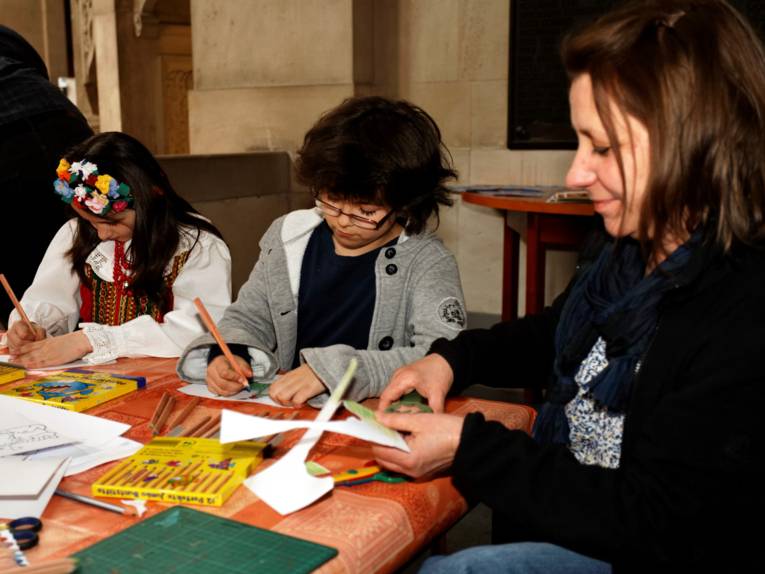 zwei Kinder, eines davon in bunter Tracht, malen mit Buntstiften auf einem niedrigen Tisch, am Rand sitzt eine erwachsene Betreuerin