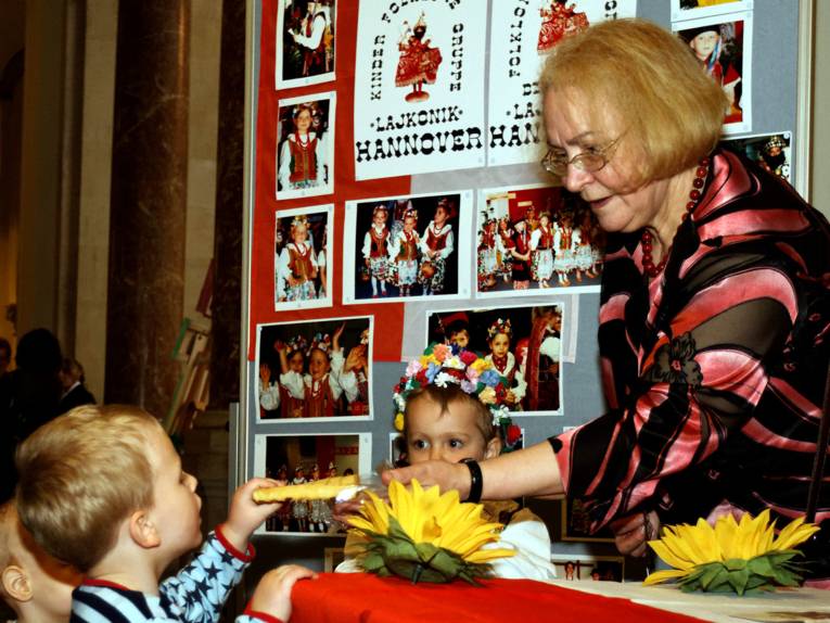 Frau am Stand der Kinderfolkloregruppe Laikonik Hannover reicht zwei Kindern im Kindergartenalter etwas