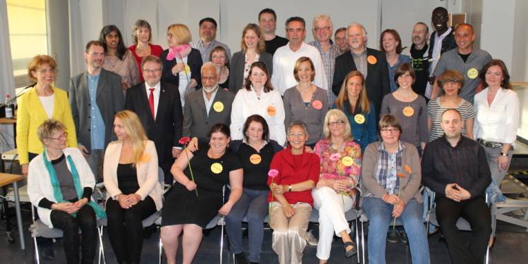 Etwa 25 Personen sitzen und stehen vor eine Bühne.