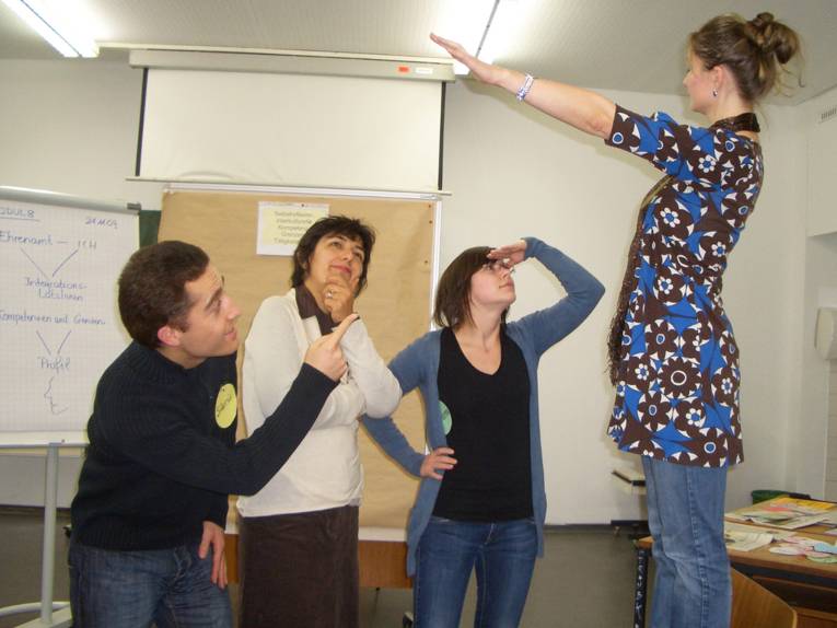 Rollenspielsituation: In einem Unterrichtsraum blicken ein Mann und ein Frau zu einer Frau auf, die mit ausgestrecktem Arm auf einem Stuhl steht, während eine dritte Frau nachdenklich wegschaut.