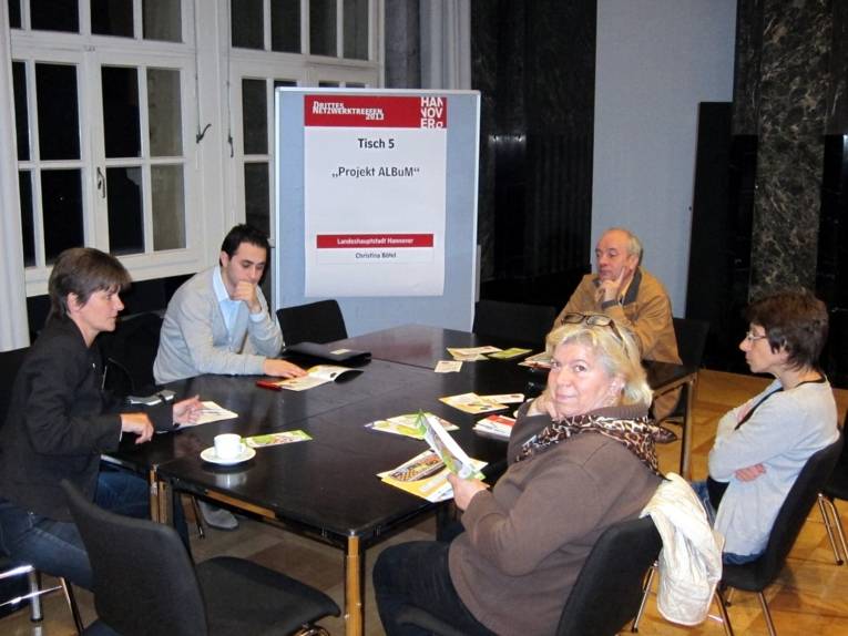 Fünf Personen sitzen an einem Infotisch im Mosaiksaal, die Referentin, Christina Bötel, sitzt links am Bildrand. Vorn dreht sich eine Teilnehmerin um und schaut direkt in die Kamera.