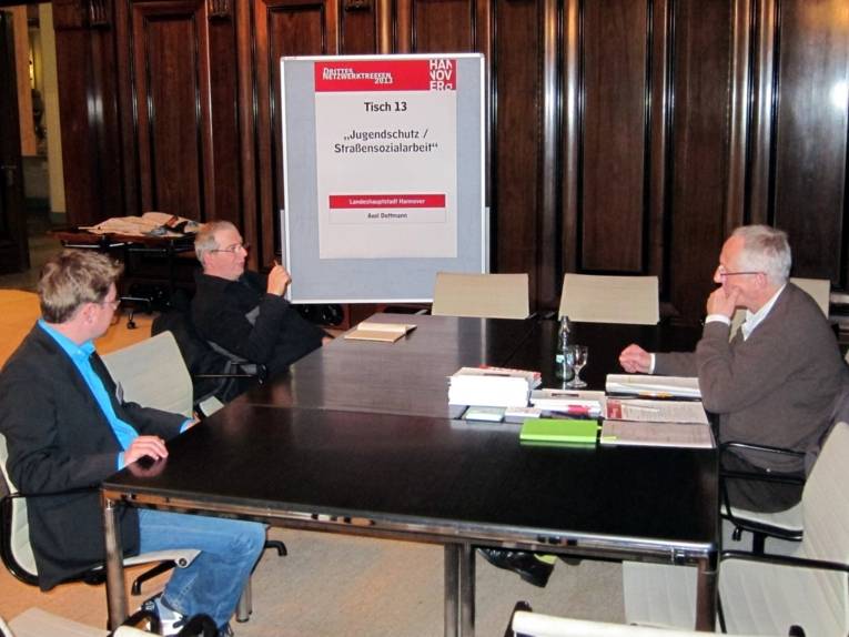 Drei Personen sitzen an einem Infotisch im Hodlersaal. Der Referent Axel Dettmann sitzt rechts im Bild, im Hintergrund zeigt ein großes Plakat auf einer Stellwand das Thema des Infotisches an.