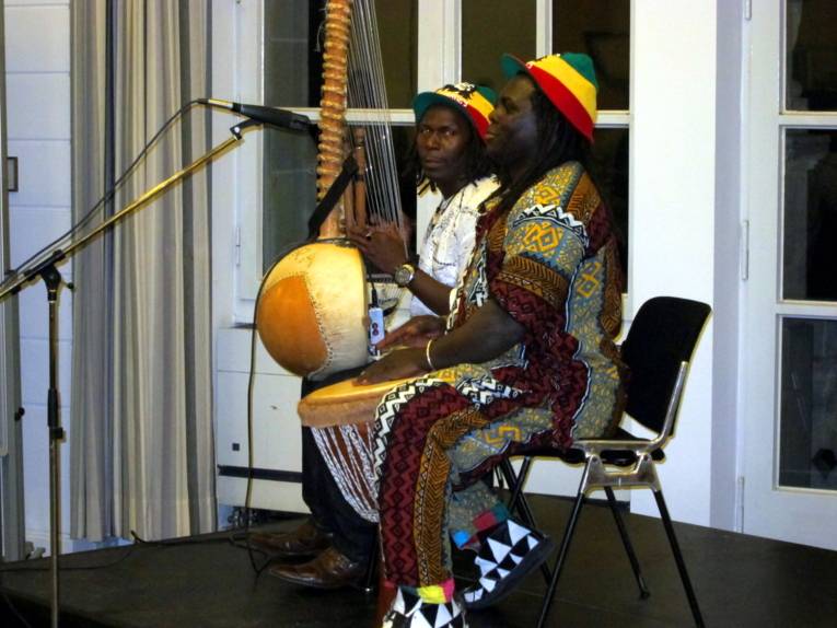 Zwei schwarzafrikanische Musiker sitzen im Gobelinsaal auf einem kleinen Podest und spielen auf Kora (eine Art Harfe) und Trommel. Vor ihnen steht ein Gesangsmikrofon.