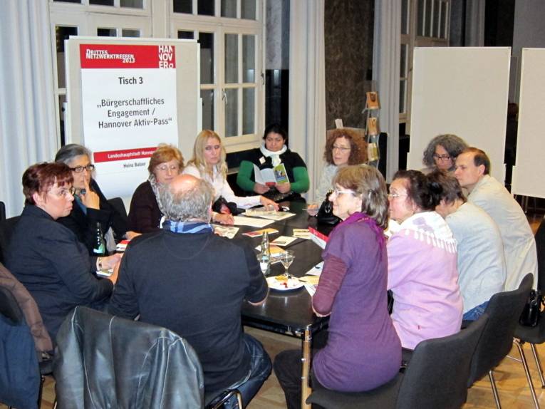 Elf Menschen sitzen um einen Tisch im Mosaiksaal und hören Heinz Balzer zu. Im Hintergrund zeigt eine Stelltafel die Themen an, die hier besprochen werden: „Bürgerschaftliches Engagement“ und „Hannover-Aktiv-Pass“.