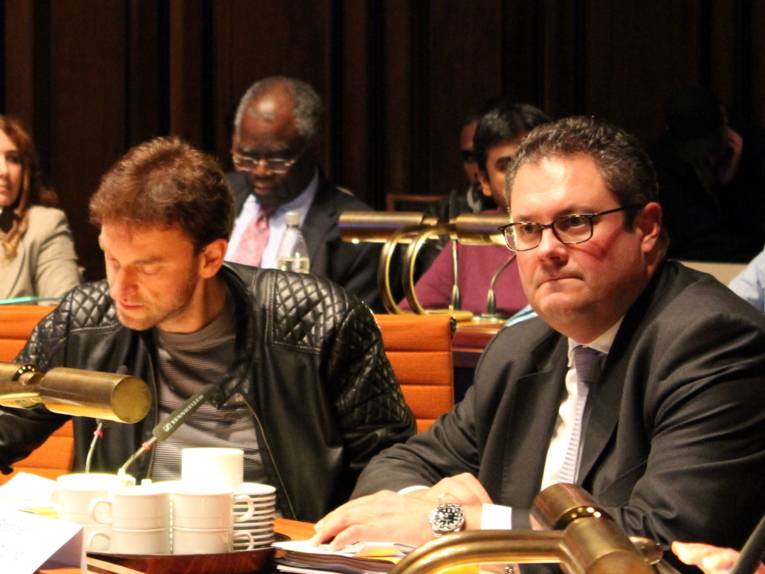 Zwei Männer sitzen im Hoflersaal hinter Pulten. Der Linke blickt auf Unterlagen, die auf seinem Tische liegen, der Rechte blickt nach vorn.