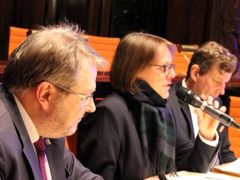 Drei Personen - zwei Männer und eine Frau - sitzen im Hodlersaal hinter Pulten. Die Frau in der Mitte spricht in ein Mikrofon, das sie in der Hand hält.