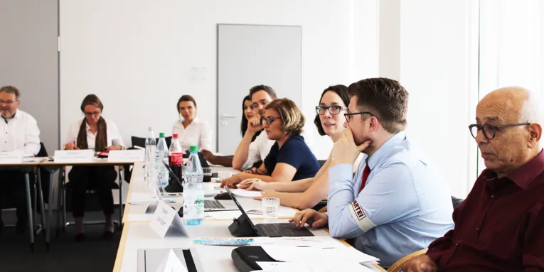 Neun Personen - fünf Frauen und vier Männer - sitzen an Tischen, die einen rechten Winkel bilden.