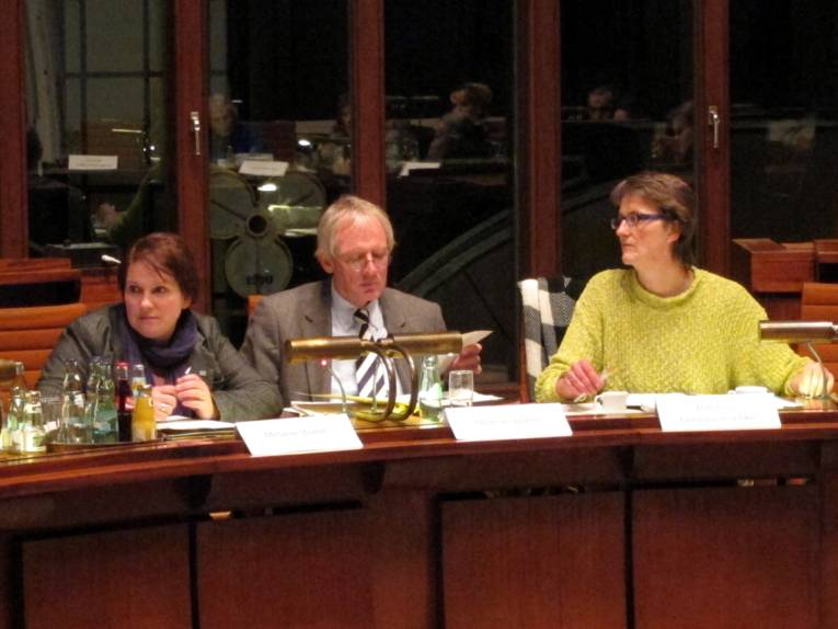 Ein Mann und zwei Frauen sitzen im Hodlersaal hinter schweren Sitzungspulten, in der dunklen Fensterfront hinter ihnen spiegeln sich die anderen Mitglieder des Ausschusses.