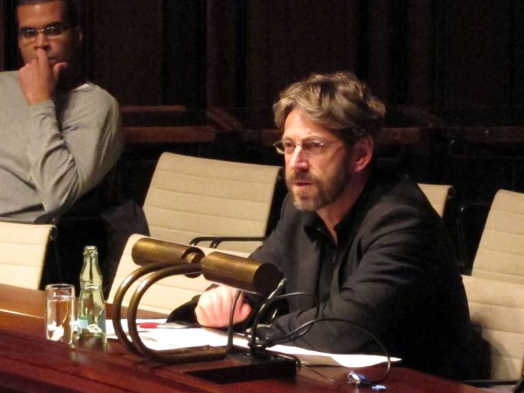 In der Mitte des Bildes sitzt eine Person mit Brille und Bart in einer Sitzreihe im Hodlersaal und spricht. Links oben im Hintergrund sitzt eine weitere Person und hört ihm zu.