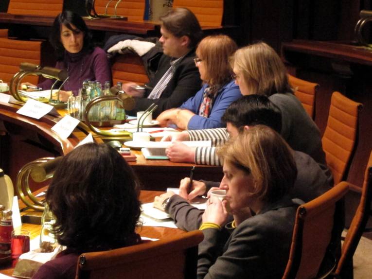 Sieben Personen sitzen an zwei gebogenen Pulten. Die vier oberen schauen auf ein Papier, das vor Kelich (auch oben im Bild) auf den Tisch liegt. Die drei anderen schauen auf Papiere, die vor ihnen auf dem Tisch liegen.