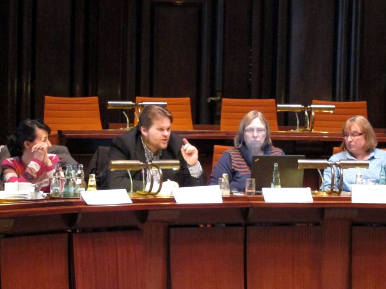 Vier Personen sitzen hinter Pulten, eine Person spricht mit erhobenem Zeigefinger. Die linke Person schaut zum Redner rechts neben ihr, die beiden rechten Personen schauen in ein aufgeklapptes Notebook auf dem Tisch vor sich.
