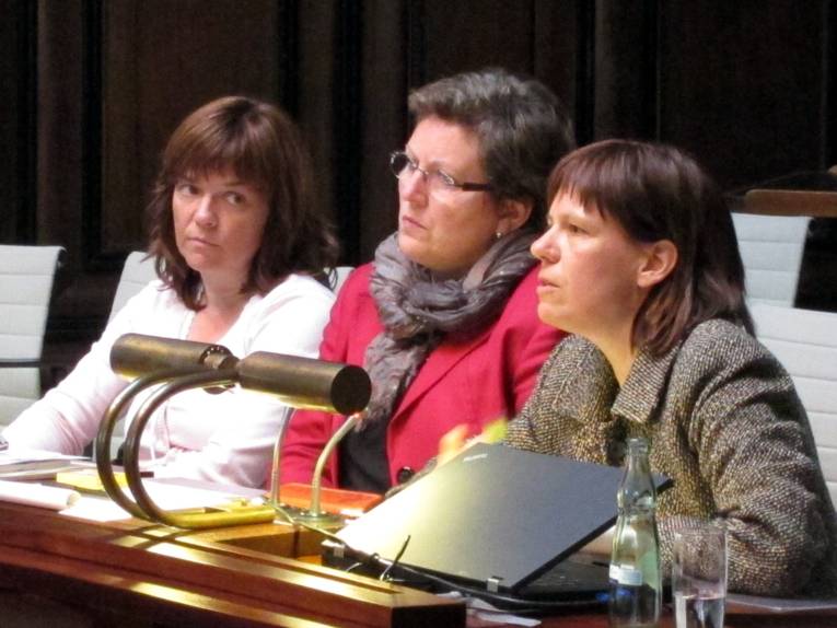 Drei Frauen sitzen nebeneinander hinter Pulten. Die Frau rechts spricht.