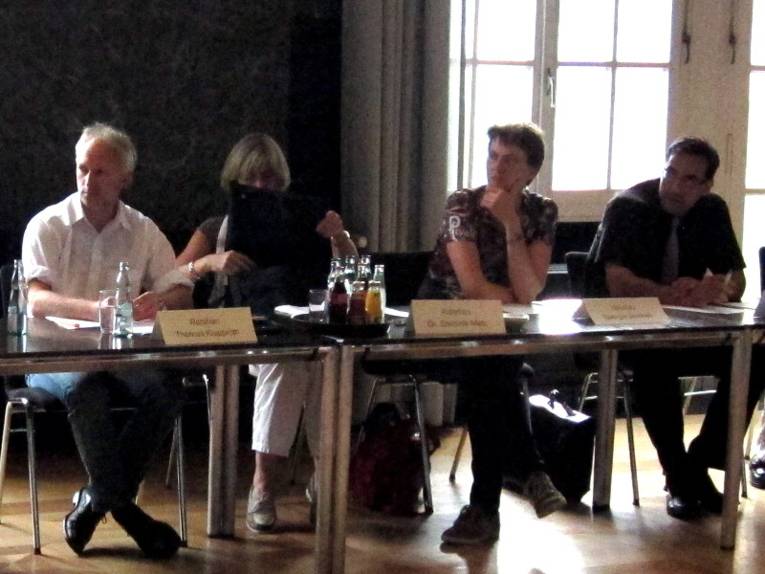 Vier Personen sitzen nebeneinander hinter Tischen. Eine Frau greift in eine Tragetasche auf ihrem Schoß.