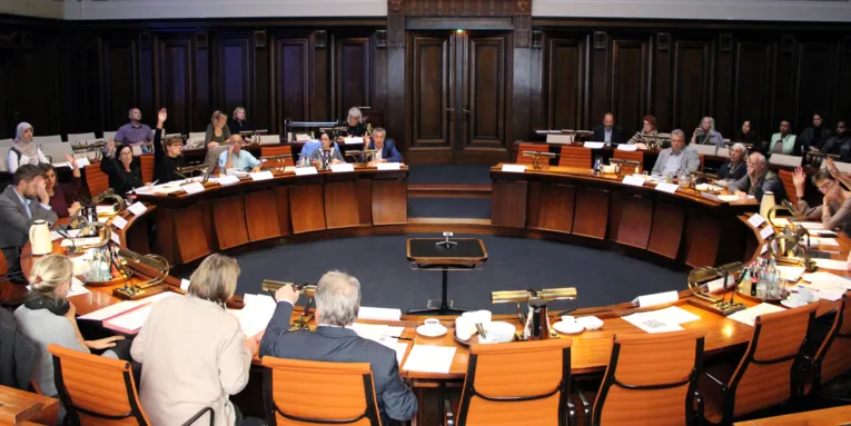 Die Ausschussmitglieder sitzen im Hodlersaal des Neuen Rathauses. EInige davon heben die Hand zur Abstimmung