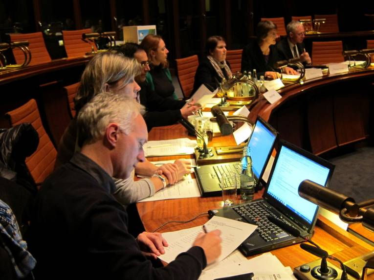 Sieben Personen sitzen im Viertelkreis im Hodlersaal und blicken entweder nach vorne, oder auf Papiere oder Notebooks auf den Pulten vor ihnen.