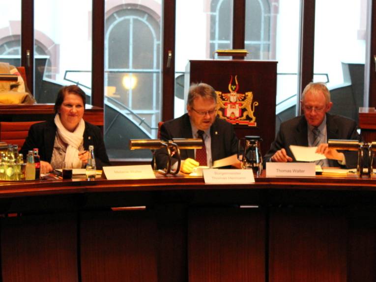 Drei Personen - eine Frau und zwei Männer - sitzen hinter Pulten. Die Frau links schaut zur Seite, die beiden Männer schauen auf Papiere, die vor ihnen auf dem Tisch liegen.