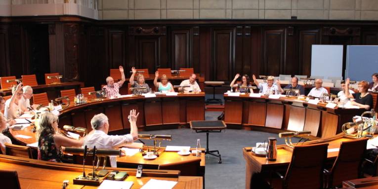 Etwa 20 Personen sitzen kreisförmig hinter Pulten im Hodlersaal im Neuen Rathaus. Die elf stimmberechtigten Mitglieder heben die Hand zur Abstimmung.