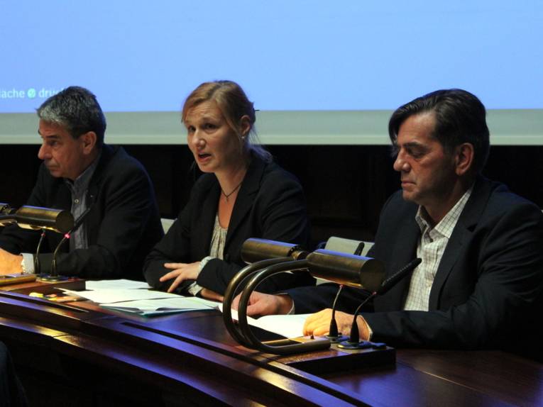 Drei Personen - zwei Männer und eine Frau sitzen im Hodlersaal hinter Pulten. Die Frau in der Mitte spricht.