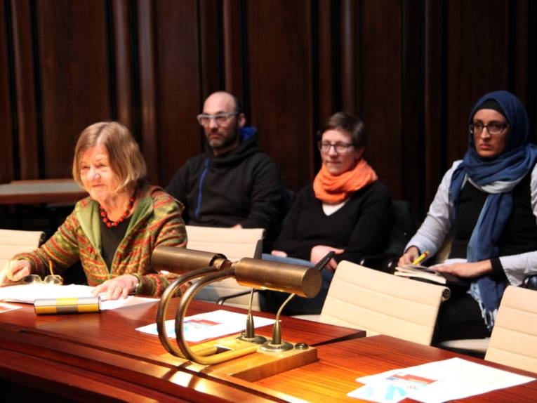 Eine Frau sitzt im Hodlersaal hinter einem Pult und spricht in ein Mikrofon. Hinter ihr sind drei weitere Personen erkennbar.