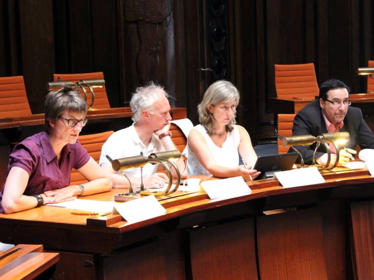 Vier Personen - zwei Frauen und zwei Männer - sitzen im Hodlersaal hinter Pulten. Die Frau links spricht in ein am Tisch befestigtes Mikrofon.